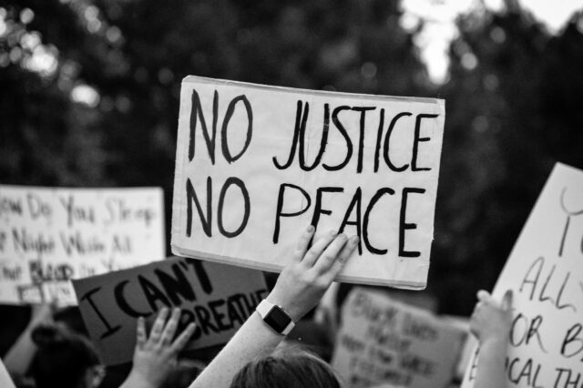 A black and white photo of protestors holding up signs. Front and center, the sign says “No justice, no peace.” Another sign in the background says “I can’t breathe.”