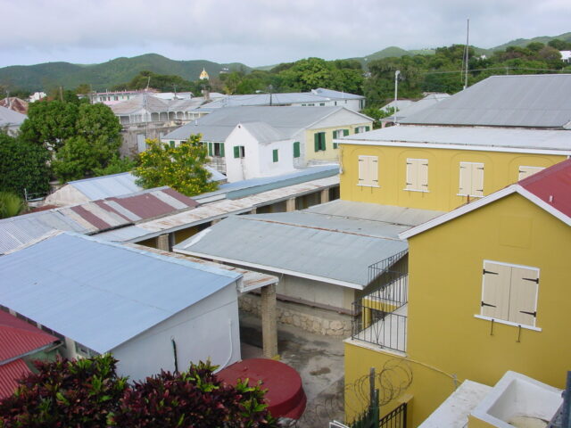Photo looks out at Frederiksted, which is downwind from the refinery. The buildings in downtown Frederiksted are charming and colorful, and there are green hills visible in the background.