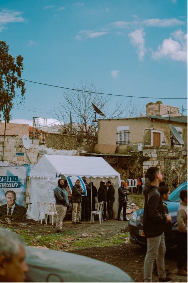 A white tent with Israeli flags on it and people inside, sitting in a yard with a stone house behind it.