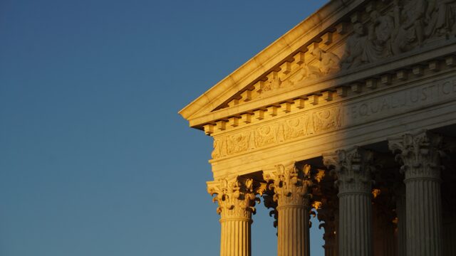 A partial façade of the Supreme Court. The sun is lighting up part of the building set against a bright blue sky.