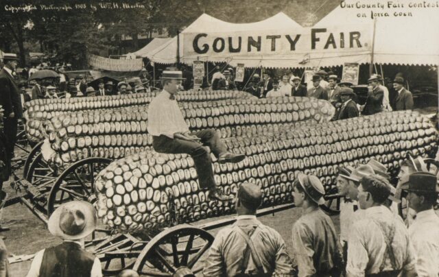 A 1908 artist’s rendering of Iowa’s corn culture: a man in a jaunty hat riding a giant ear of corn in front of a big white tent marked “County Fair.”