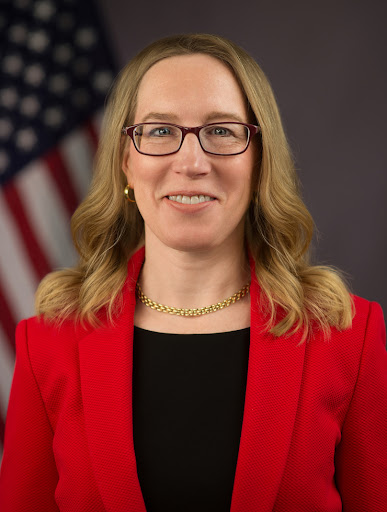 Official photo of Hester Peirce smiling, wearing a red blazer with a black shirt, with an American flag in the background