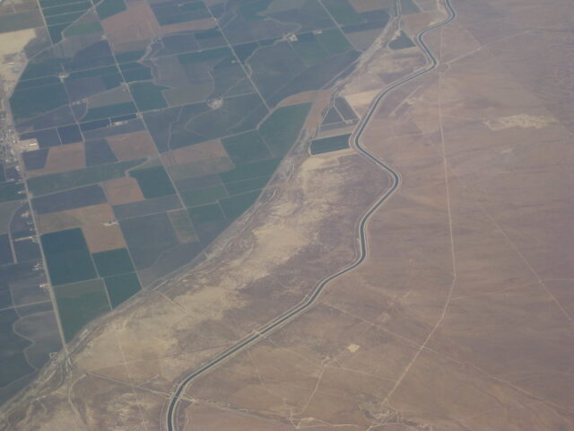 Aerial view of a landscape. The top and left half of the image shows lots of green squares of farmland. There are some brown squares, but most are a dark green. The bottom and right half of the image shows a barren, empty brown landscape. There is a road that follows the boundary between the empty land and farmland.