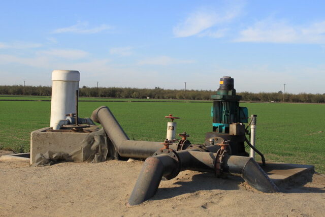 A structure sits atop sand-colored ground right on the edge of a bright green field. The structure consists of thick, brown pipes sticking out of the ground and attached to a pumping machine. A white tube-like structure sits right next to the pipes, atop a cement block.
