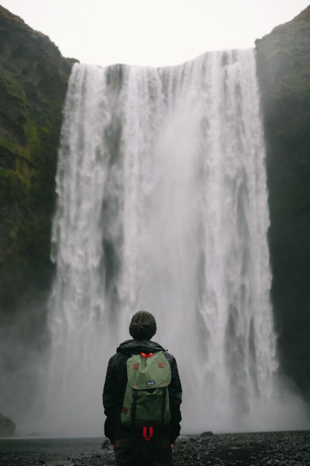 A person stands with their back to the camera looking at a large waterfall while wearing a Patagonia backpack.