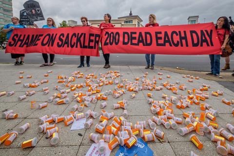 Protestors with a sign reading “shame on Sackler” stand surrounded by empty pill bottles.