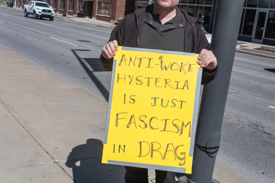 Person holds a sign that says “Anti-Woke Hysteria Is Just Fascism In Drag.” 
