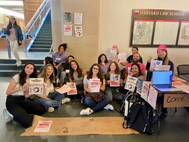About a dozen of Harvard Law students sit on the floor of an academic building as they hold signs promoting reproductive justice programming at Harvard Law School 