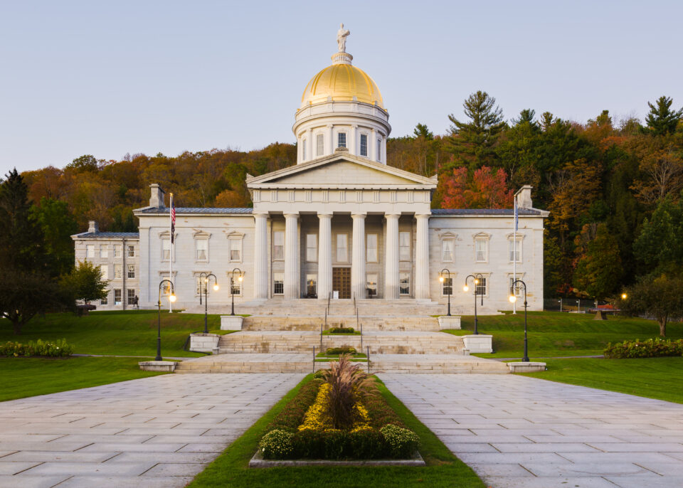 The Vermont Statehouse, a stately, white building with columns and a gold-domed roof on an early autumn day.