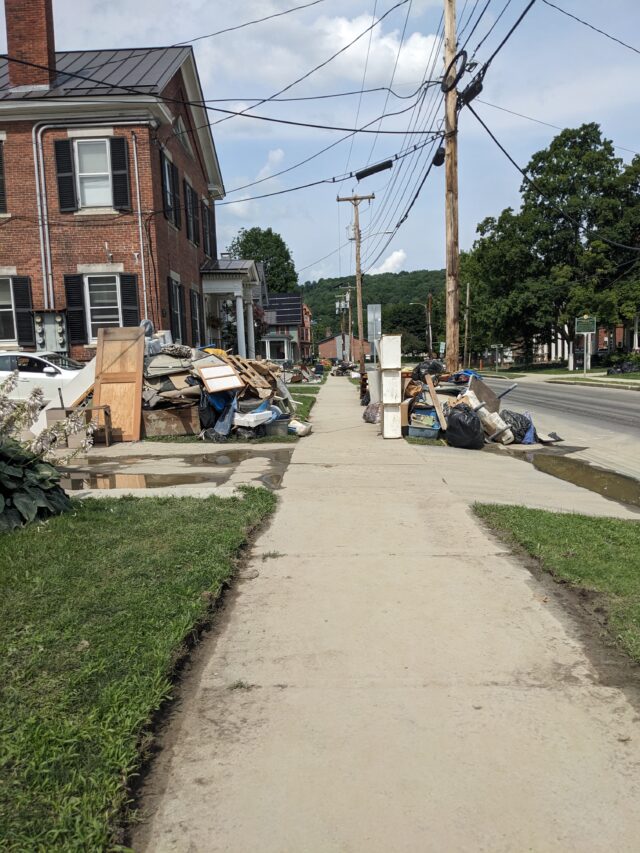 On a residential street, a large pile of household furniture and items are piled up on the lawn and next to the sidewalk. 