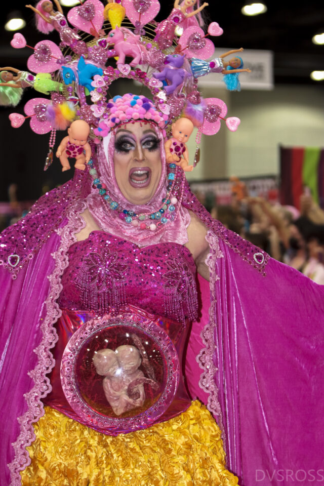 A person in female drag wears a pink headpiece and pink bustier and shawl with twin embryos displayed through a clear womb.