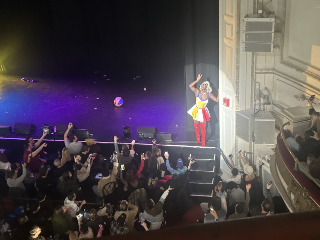 A person in female drag dressed as Katy Perry in her “California Girls” music video performs for an audience. A beach ball and dollar bills lay on the stage in the background.