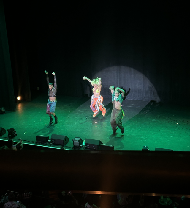 A person in female drag dressed in orange baggy pants and a neon yellow bra jumps up between two male dancers on a stage lit in bright green.