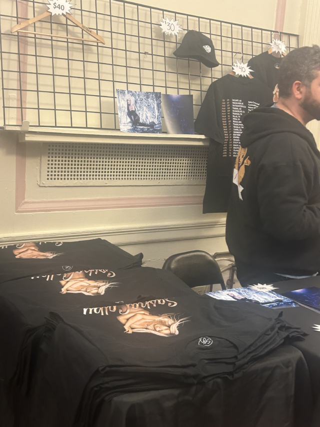 A merchandise table at Sahsa Colby’s “Stripped” tour. The shirt listed for sale at $40 appears to be sold out, while other items can be seen throughout the background. A pile of black t-shirts lay on the table in the foreground.