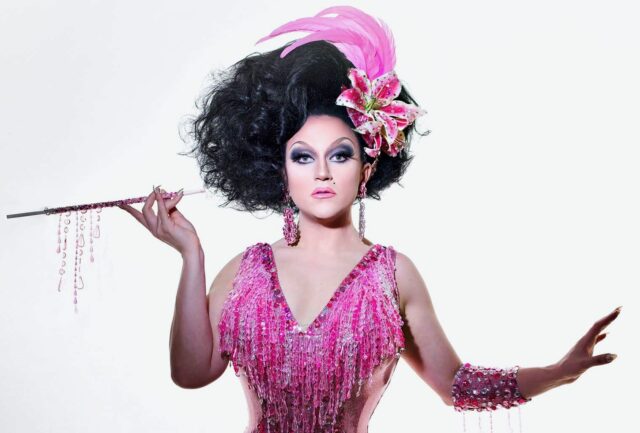 A person in female drag dressed in a pink fringed outfit and short black hair holds a cigarette in an elongated cigarette holder.