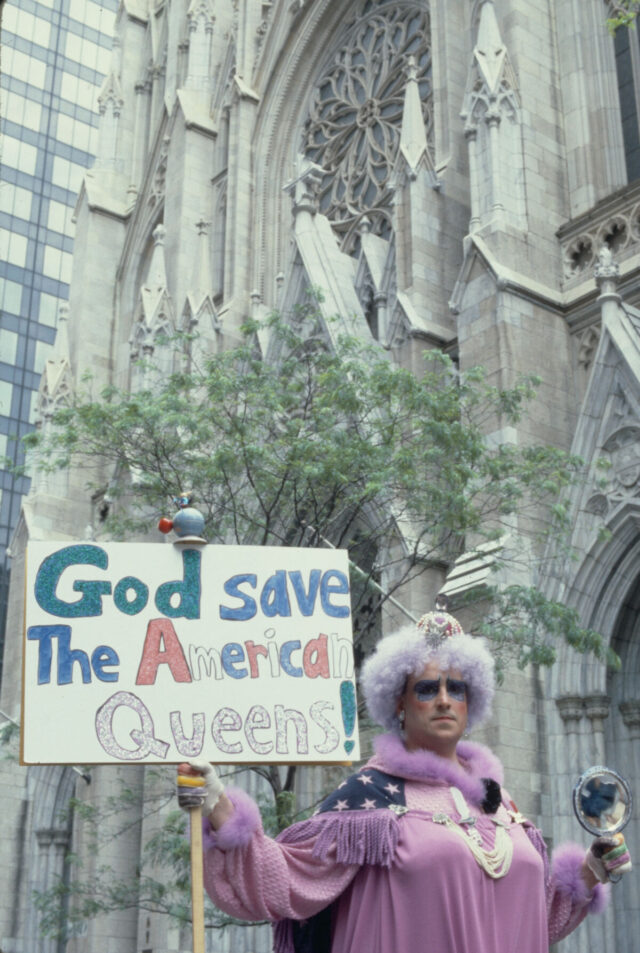 A person in women’s dress holds a sign that reads “God save the American queens!” while standing in front of a church. 