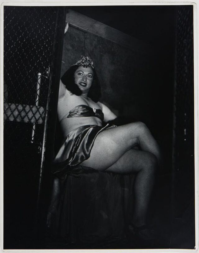 A person in women’s dress smiles from the inside of a police vehicle.