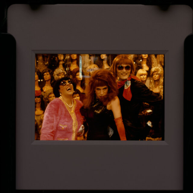 Three people in female drag pose in front of mannequin heads in wigs.