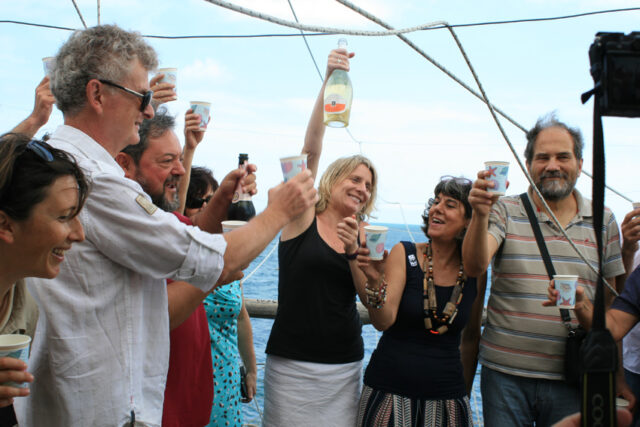  Maria D’Orsogna, surrounded by a crowd of people, smiles while holding up a bottle of sparkling wine.