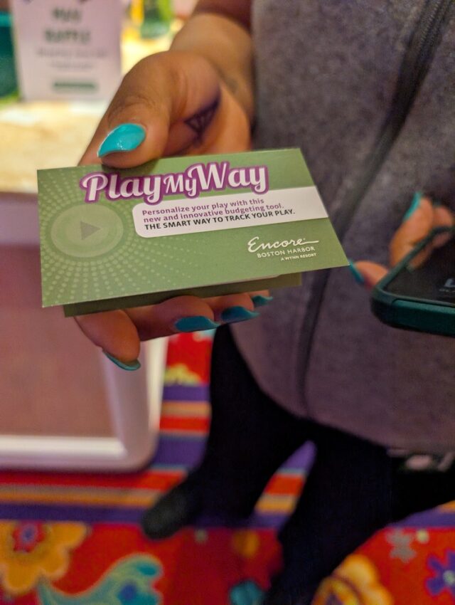 A manicured hand with teal painted nails holds a business card that says “Play My Way.” 