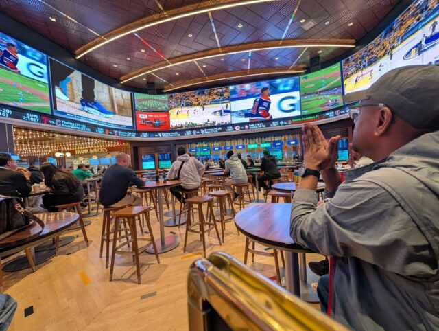 Sports fans sit on barstools with their back to the camera. They look up at multiple sporting events on giant televisions. 
