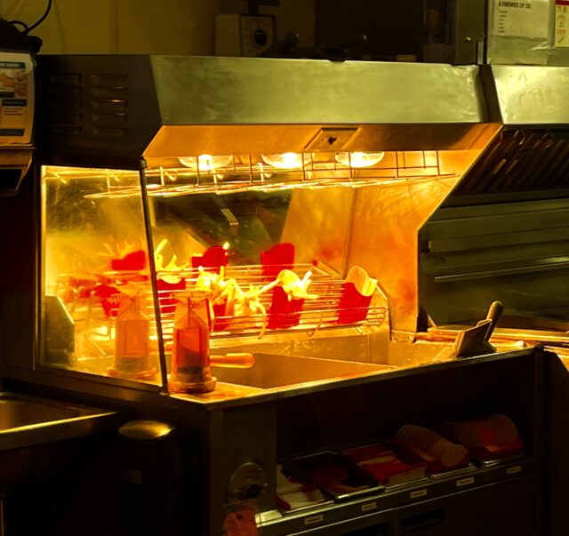McDonald’s heated fry station under yellow heat lamps, with several red fry cartons holding cooked fries on a wire shelf, salt shakers on the counter’s edge, and stainless-steel kitchen equipment in the background. 