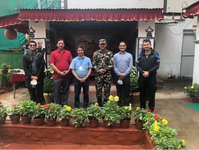 A group of Nepal men pose in front of a flowerbed at an army base, including an army officer, four activists, and a local priest.