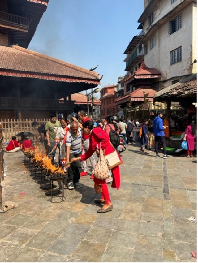Nepali men and women, one in traditional red dress, use sticks to tend to about a dozen small, blazing coal fires in pots raised up on stands in the square surrounded by temples.