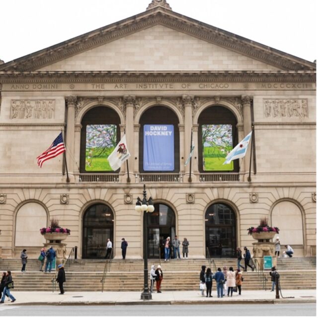 The front of the Art Institute of Chicago’s tan marble building with the flags of the U.S., Chicago, and Illinois blowing in the wind, visitors standing on the steps and on the street, and three banners for an exhibit at the time for David Hockney.