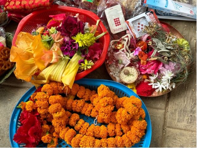 Three trays sit on the floor, one filled with circular garlands of orange marigolds to put around the necks of statues of deities, one with assorted flowers to decorate their shrines, and one with red vermillion powder, rice, and red circular stickers to decorate and/or paint their faces.
