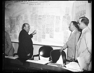 Howard Hopson gesturing toward an intricate diagram of AG&E’s corporate form as two senators look on.