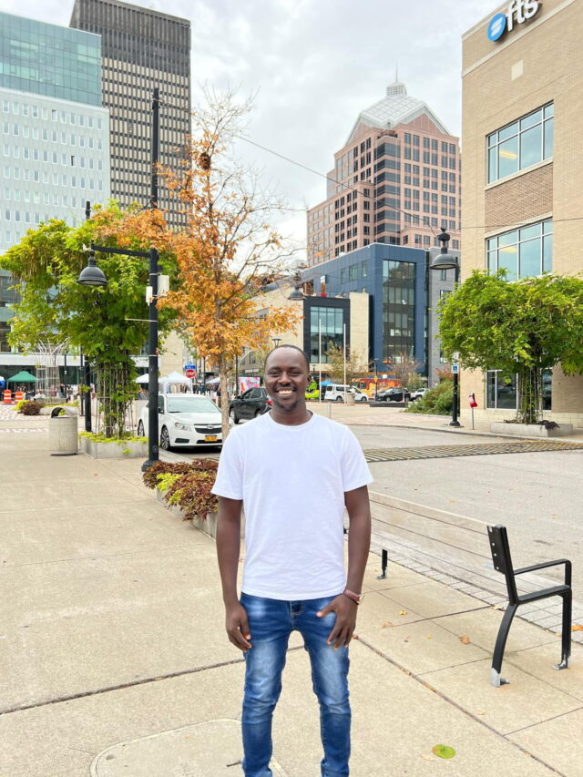 Issakh stands on a sidewalk, smiling in front of office buildings.