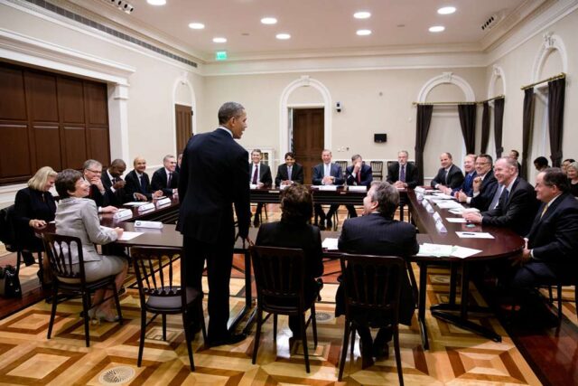 President Obama stands and addresses a group of Business Roundtable CEOs, who sit in a circle and smile.