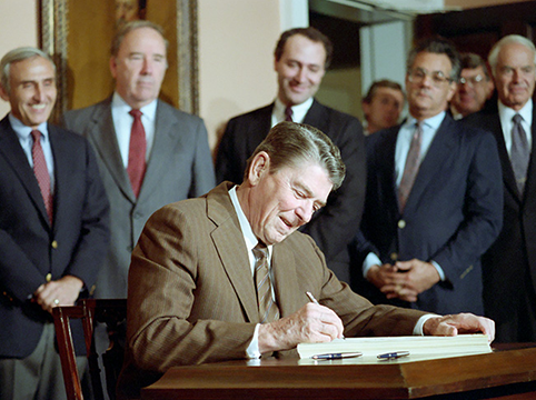 President Reagan, wearing a brown suit, signs IRCA into law, smiling, as other politicians look on in the background.
