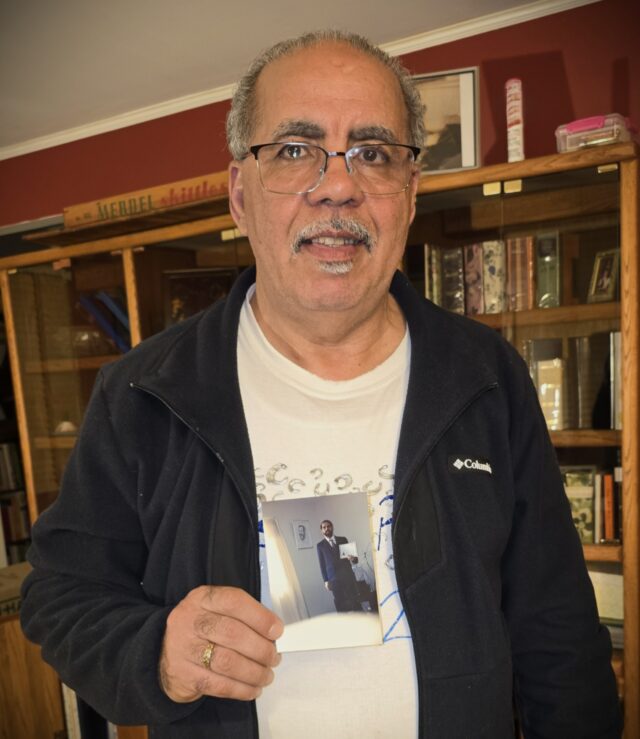 My father, wearing a fleece sweater and a t-shirt, smiles slightly, holding the photo of him from 1998 against his chest.