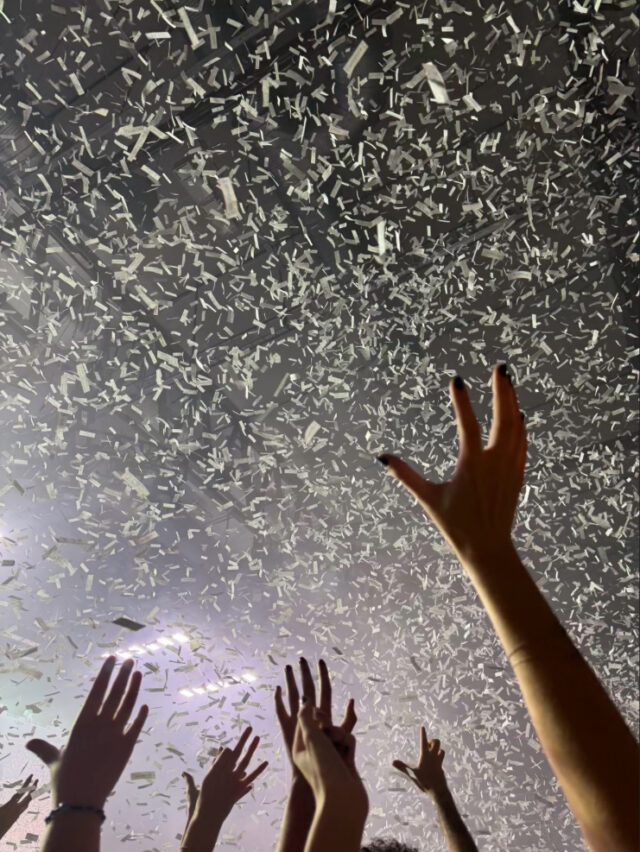 Raised hands in a crowd at a concert with white confetti falling from above under stage lighting.